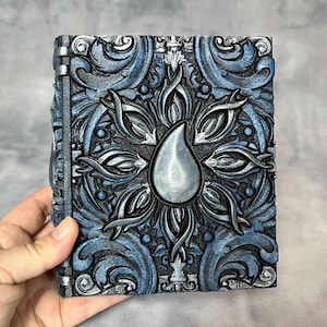 May include: An intricately designed square box, featuring a raised floral pattern in blue and silver. The central element is a teardrop shape. The box has a textured finish and is held in a hand.