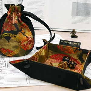 Peut inclure: Un plateau à dés en tissu noir avec un motif de dragon et de nœud celtique. Le plateau est ouvert et contient des dés. Le plateau porte une étiquette indiquant "Geeky Tendencies".