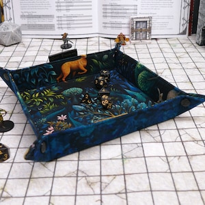 Peut inclure: Un plateau de dés en tissu avec un motif forestier dans les tons de bleu, vert et rose. Le plateau est plié et boutonné aux coins. Des dés et des figurines miniatures sont à l'intérieur. Une petite pancarte indique "Great Fortress".