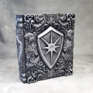 May include: A silver-coloured, rectangular box with an embossed design. The front features a shield with a starburst design. The box has ornate scrollwork and a book-like spine, suggesting a decorative storage item or container.