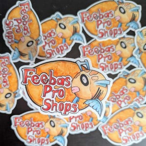 May include: A collection of stickers featuring a cartoon fish with the text "Feebas Pro Shops" in red and white. The fish is orange, with blue fins and a pink tongue. The stickers are on a black surface.