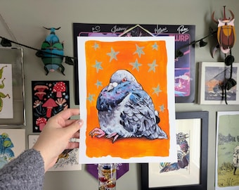 May include: A watercolor print of a plump pigeon with red eyes, set against an orange background with teal stars. The bird is depicted in shades of gray, blue, and white. The artwork is held by a person.