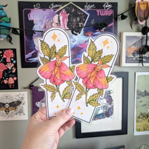 May include: Two clear, plastic-covered stickers featuring a pink and orange moth design with green leaves and stems. The moth stickers have a white border and are decorated with yellow star accents.