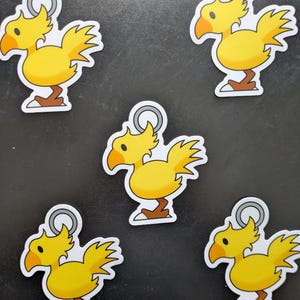 May include: Five yellow Chocobo stickers with orange beaks and brown feet are displayed. Each sticker has a silver loop at the top. The stickers are on a dark gray surface.