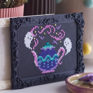 May include: Two decorative frames, one cream and one black, are displayed. The black frame showcases a cross-stitch design of a teapot with ghosts and decorative elements in pink, teal, and white. A small dish with colourful thread is in the foreground.