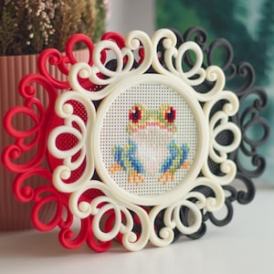 Pode incluir: Um bordado em ponto cruz circular apresentando um sapo colorido com olhos vermelhos, membros azuis e pés amarelos. O bordado está inserido numa moldura decorativa de plástico creme com detalhes em filigrana, sobreposta a molduras vermelha e preta.