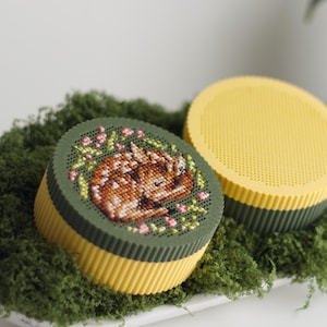 Puede incluir: Dos cajas redondas decorativas. Una presenta un diseño de punto de cruz de un ciervo durmiente rodeado de flores rosas y hojas verdes. La otra es amarilla con una base verde. Ambas descansan sobre musgo verde.