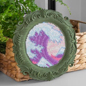 Pode incluir: Um bordado em ponto cruz circular apresentando um padrão de onda estilizado em tons de roxo, rosa e azul claro. O bordado está inserido numa moldura verde-oliva mate ornamentada com volutas intrincadas e detalhes em forma de folha.