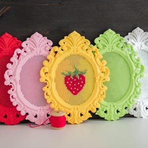 Five baroque oval cross stitch frames in red, pink, yellow, green and white lined up on a shelf, the yellow one with a stitched strawberry design, shown with a spool of red thread. Stitchable frames printed from the STL file.