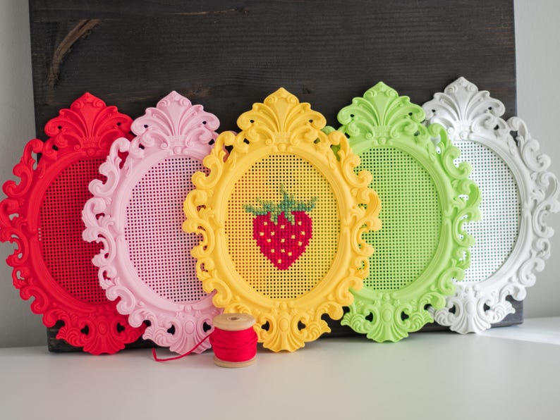 Five baroque oval cross stitch frames in red, pink, yellow, green and white lined up on a shelf, the yellow one with a stitched strawberry design, shown with a spool of red thread. Stitchable frames printed from the STL file.