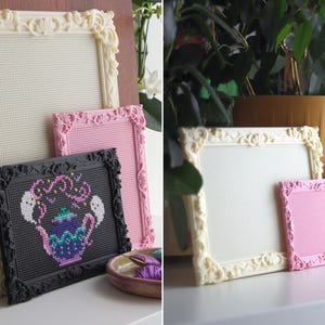 May include: A collection of rectangular frames with ornate, raised scrollwork borders in cream, pink, and black. The frames feature a grid-like interior surface. One black frame contains a cross-stitch design of a purple teapot with two white ghosts.