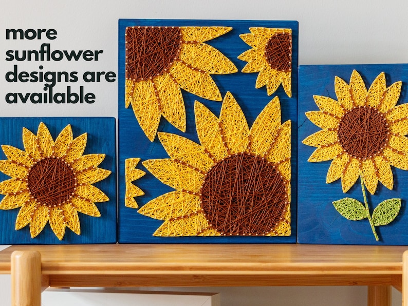 以下が含まれることがあります： 青い木製の壁飾り3点セット。糸で作ったひまわりが描かれています。一番大きいものはひまわりが2つ、他の2つはそれぞれひまわりが1つ描かれています。