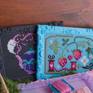 May include: Three rectangular plastic frames with ornate borders containing cross-stitch designs. One black frame features a purple teapot with a ghost, a blue frame displays strawberries and sewing spools, and a light purple frame holds pink thread.