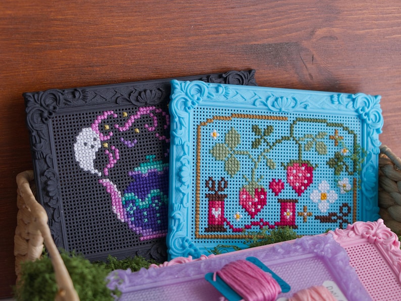 May include: Three rectangular plastic frames with ornate borders containing cross-stitch designs. One black frame features a purple teapot with a ghost, a blue frame displays strawberries and sewing spools, and a light purple frame holds pink thread.