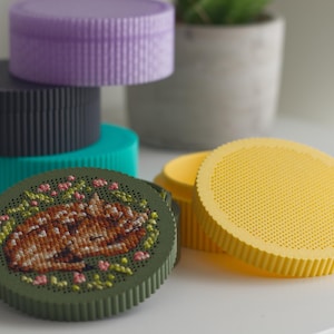 Round cross stitch box STL file — 3D printed stitchable boxes in purple, grey, teal, green and yellow PLA, with sleeping fawn embroidery on the green lid and a blank yellow perforated lid
