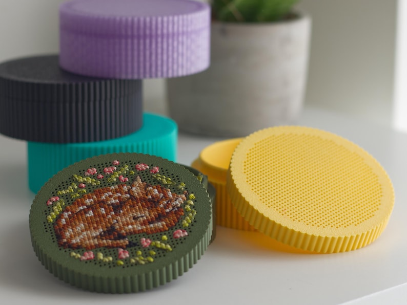 Round cross stitch box STL file — 3D printed stitchable boxes in purple, grey, teal, green and yellow PLA, with sleeping fawn embroidery on the green lid and a blank yellow perforated lid
