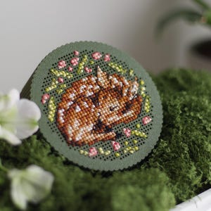 Round cross stitch box STL file — 3D printed lid with sleeping fawn and floral wreath embroidery, shown in green PLA on moss with white blossoms