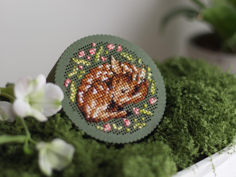 Round cross stitch box STL file — 3D printed lid with sleeping fawn and floral wreath embroidery, shown in green PLA on moss with white blossoms