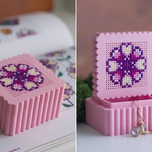 3D printed pink cross stitch box lid with flower embroidery in progress next to a pattern book, and stacked finished pink and green stitchable boxes