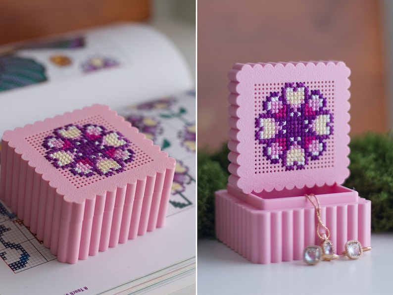 3D printed pink cross stitch box lid with flower embroidery in progress next to a pattern book, and stacked finished pink and green stitchable boxes
