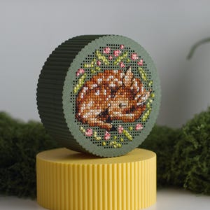 Round cross stitch box STL file — 3D printed green keepsake box with sleeping fawn and floral wreath embroidery on the lid, displayed on a yellow box