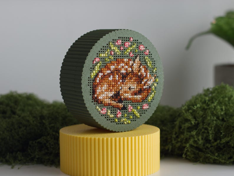 Round cross stitch box STL file — 3D printed green keepsake box with sleeping fawn and floral wreath embroidery on the lid, displayed on a yellow box