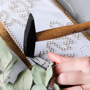 May include: A person uses a hammer and pliers to secure gold-colored tacks into a wooden frame with a white surface. The tacks are arranged in a pattern.