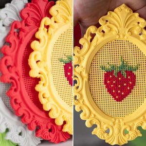 Baroque oval cross stitch frames in white, red, yellow and green, with a close-up of a yellow frame held in hand showing a finished strawberry design. Stitchable frames printed from the STL file.