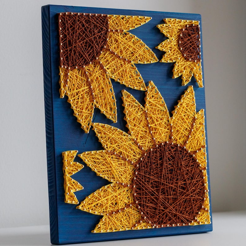 以下が含まれることがあります： 青い木製の板に、黄色と茶色の糸で作ったひまわりが3つ描かれている。ひまわりは、中央に一番大きなひまわりがくるように、集まって配置されている。