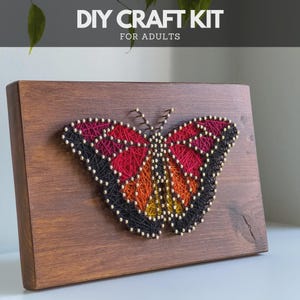 Könnte beinhalten: DIY-Bastelset für Erwachsene mit einem Schmetterlings-String-Art-Design. Der Schmetterling besteht aus rotem, orangefarbenem, gelbem und schwarzem Faden auf einer gebeizten Holzplatte. Der Text "DIY CRAFT KIT FOR ADULTS" steht oben.