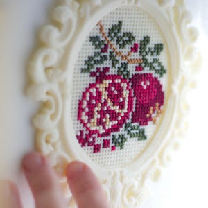 Cream baroque oval cross stitch frame with a finished pink pomegranate floral design stitched into the grid, held up to the light. Stitchable frame printed from the STL file.