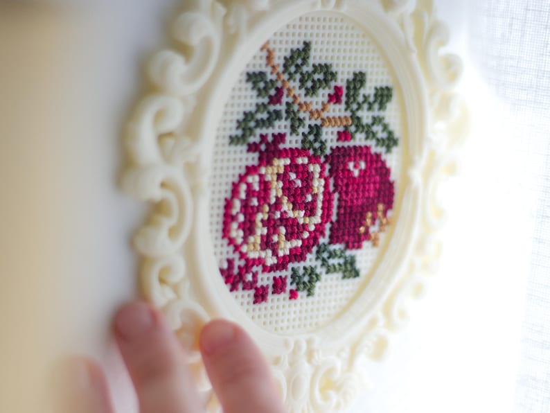 Cream baroque oval cross stitch frame with a finished pink pomegranate floral design stitched into the grid, held up to the light. Stitchable frame printed from the STL file.