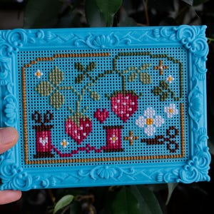 May include: A cross-stitch embroidery piece featuring strawberries, flowers, and sewing spools in a bright blue plastic frame. The design includes red strawberries, green leaves, white blossoms, and two pink spools of thread connected by a red line.