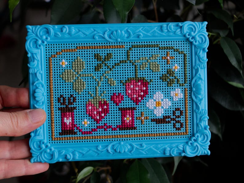 May include: A cross-stitch embroidery piece featuring strawberries, flowers, and sewing spools in a bright blue plastic frame. The design includes red strawberries, green leaves, white blossoms, and two pink spools of thread connected by a red line.