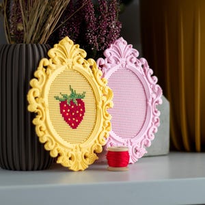 Yellow and pink baroque oval cross stitch frames on a shelf, the yellow one with a stitched strawberry design and the pink one blank, shown with a spool of red thread. Stitchable frames printed from the STL file.