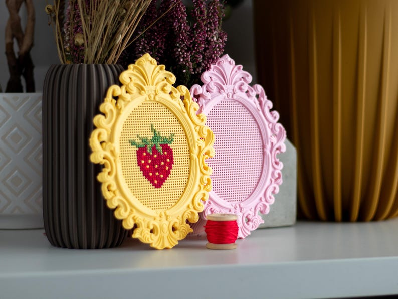 Yellow and pink baroque oval cross stitch frames on a shelf, the yellow one with a stitched strawberry design and the pink one blank, shown with a spool of red thread. Stitchable frames printed from the STL file.