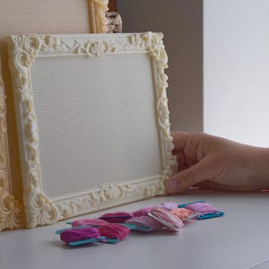 May include: Two cream-colored, rectangular embroidery frames with ornate, raised floral and lattice detailing along the borders. The frames contain a white, grid-patterned fabric mesh. Several small, flat spools of pink, purple, and peach embroidery thread.