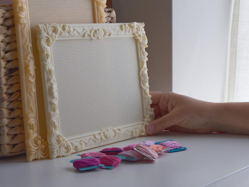 May include: Two cream-colored, rectangular embroidery frames with ornate, raised floral and lattice detailing along the borders. The frames contain a white, grid-patterned fabric mesh. Several small, flat spools of pink, purple, and peach embroidery thread.