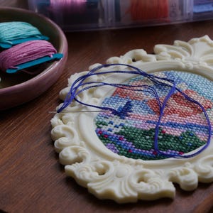 Cream baroque oval cross stitch frame with a colourful landscape design in progress, a threaded needle resting on the grid, shown with a dish of embroidery floss. Stitchable frame printed from the STL file.
