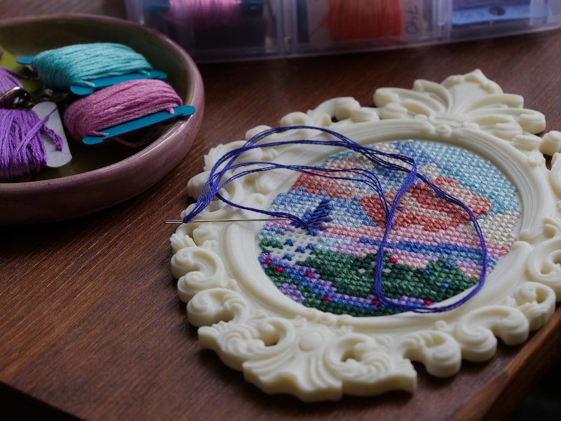 Cream baroque oval cross stitch frame with a colourful landscape design in progress, a threaded needle resting on the grid, shown with a dish of embroidery floss. Stitchable frame printed from the STL file.