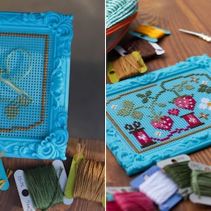 May include: A bright turquoise plastic frame containing a cross-stitch pattern of strawberries, green leaves, and white flowers. The embroidery is worked on a blue perforated grid. Several spools of embroidery floss in various colors are nearby.