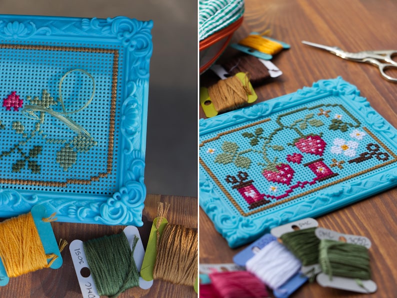 May include: A bright turquoise plastic frame containing a cross-stitch pattern of strawberries, green leaves, and white flowers. The embroidery is worked on a blue perforated grid. Several spools of embroidery floss in various colors are nearby.