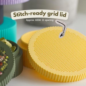 Round cross stitch box STL file — blank stitch-ready perforated lid with AIDA 11 spacing grid, shown in yellow PLA next to a green box with sleeping fawn embroidery