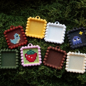 Puede incluir: Una colección de ocho marcos cuadrados perforados con bordes festoneados en varios colores, incluyendo amarillo, blanco, negro, rosa, marrón y verde. Algunos marcos contienen diseños de punto de cruz de un pájaro azul, una fresa y un fantasma morado.