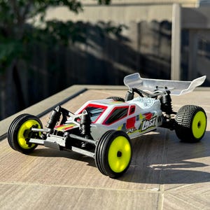 May include: A white, red and yellow radio-controlled off-road buggy with a clear plastic wing. The buggy has the word "Lost" printed on the side.