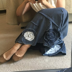 Op de afbeelding: Een vrouw draagt een wit, mouwloos topje en een blauwe en witte stippenrok met witte geborduurde bloemen. Ze zit op een beige kleed met haar benen gekruist en draagt ​​bruine ballerina's.