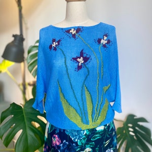 Puede incluir: Una blusa azul claro con un diseño floral. La blusa tiene mangas largas y cuello redondo, con un patrón de flores moradas y blancas con tallos verdes. La parte inferior del maniquí lleva una falda con estampado floral.