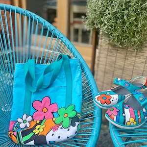 May include: A blue tote bag with a colorful floral design. The bag has two teal straps and is sitting on a blue chair.