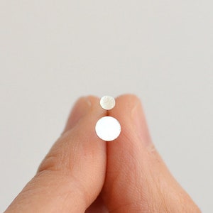 May include: Two white pearl earrings, one larger than the other, held between two fingers.