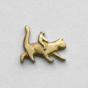 Könnte beinhalten: Eine goldfarbene Metallstifte mit einem stilisierten Bild einer Katze, auf der eine Person reitet.